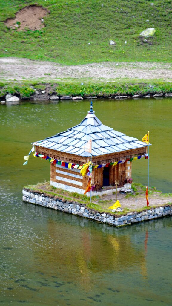 A traditional wooden hut sits on a serene lake, surrounded by lush greenery, under a summer sky.